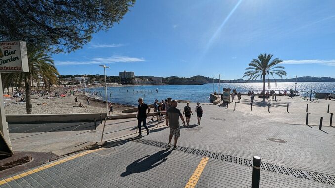 Zicht op het strand en de boulevard van Peguera op Mallorca, met zonaanbidders, palmbomen en helderblauwe zee onder een stralende zon.